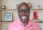 A man smiles during a video call, wearing a pink shirt and glasses, with shelves and a 'Mindful Techie' sign in the background.