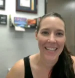 Portrait of a smiling woman with dark hair tied back, wearing a black sleeveless top, in an office with framed pictures on the wall.