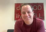 Smiling man in a maroon shirt with a red sign behind him in an indoor setting.