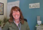 Smiling woman wearing headphones on a video call, with a teal wall and a 'LADS' sign in the background.