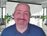 Smiling man with gray hair and beard in a blue polo shirt, standing in a modern office with a 'simple' sign in the background.