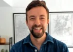 Smiling man with short brown hair and beard, wearing a blue denim shirt, indoors with framed maps in the background.