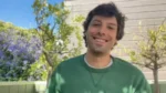 Man smiling in a green shirt outdoors with purple flowering bushes and a wooden fence behind.