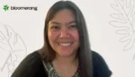 Smiling woman with long dark hair wearing a black top, in front of a white background with plant illustrations and a Blooming/ Bloomerang logo in the top-left.