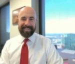 Smiling man in a white shirt and red tie sits in an office with windows behind him.