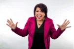 Cheerful woman in a magenta blazer stretches arms wide in a welcoming pose against a light background.