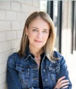 Portrait of a smiling woman with blonde hair in a denim jacket, standing against a light brick wall, arms crossed.