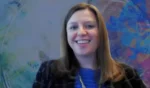 Smiling woman on a video call, wearing a blue top and dark blazer, with a colorful mural in the background.