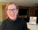 Smiling woman with red hair and glasses speaks to camera at a fundraiser, with a fundraising sign in the background.