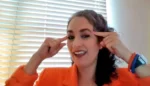 Woman in an orange blazer smiling and pointing to her temples with both index fingers to indicate thinking or concentration.
