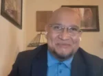 Man with glasses in a blue shirt and dark blazer speaks on a video call, wearing white wireless earbuds, with framed art and a lamp in the background.