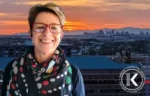 Smiling woman with short blonde hair and red glasses, wearing a colorful scarf, outdoors at sunset with a city skyline; circular logo bottom-right.