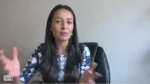 Woman with long dark hair in a blue plaid top sits in a chair and gestures with her hands while speaking to the camera.