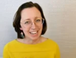 Smiling woman with short dark hair and round glasses wearing a bright yellow top, posing against a neutral patterned background.