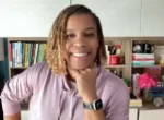 Smiling woman with braided hair in a lavender hoodie rests her chin on her hand, with a bookshelf behind her in a home office.