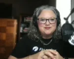 Woman with gray hair and glasses sits at a desk in a home recording setup, speaking into a microphone.