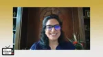 Smiling woman with dark hair and glasses on a video call, wood-paneled backdrop and a plant visible in the background.
