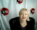 Smiling woman with blonde hair and red lipstick sits in front of a Charity Watch backdrop with red logos behind her (video call scenario).