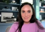 Woman with long dark hair in a pink sweater looks at the camera during a video call in a blue-toned room.