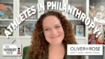 Smiling woman with curly hair in a studio, background shelves, curved banner text says 'Athletes in Philanthropy' with 'The Nonprofit Show' and 'Oliver Rose' logos.