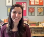 Smiling woman with long dark hair wearing a purple top, in a classroom with colorful framed art on the wall.