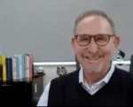Smiling man in a blue shirt and black vest wearing glasses in an office, with books on a shelf behind him.
