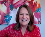 Smiling woman in a red patterned blouse speaks during a video call with a bright abstract painting in the background.