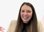 Smiling woman with long brown hair wearing a beige blazer, on a video call against a bright white background.