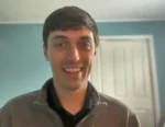 Smiling man with short dark hair wearing a gray zip-up jacket, indoors with blue walls and a white door in the background.