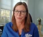 Woman with shoulder-length brown hair and glasses wearing a blue blouse, speaking on a video call.