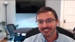 Man with glasses smiling during a video call in a bright home office setting, with a desk and plants in the background.