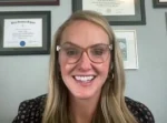 Smiling blonde woman with glasses sits in front of framed diplomas on the wall, in a professional setting.