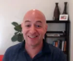 Smiling bald man in a dark polo shirt with a plant and bookshelf in the background, appearing in an office setting.