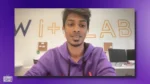 Young man in a purple hoodie speaks to the camera in a bright room with large orange letters on the wall behind him.