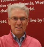 Portrait of a smiling older man with gray hair and glasses, wearing a pink zip-up sweater over a blue collared shirt, standing in front of a red background with white text.