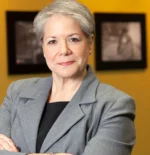 Professional woman with gray hair in a gray blazer, arms crossed, standing in an office setting with a yellow wall behind her.