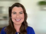 Smiling woman with long brown hair wearing a blue top, indoors with a softly blurred background.