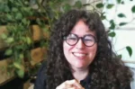 Smiling woman with curly dark hair and round glasses, in a plant-filled indoor setting.