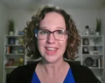 Woman with curly hair and glasses smiling at the camera in a home office setting.