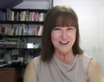Woman with shoulder-length brown hair smiling at the camera, indoors with bookshelves in the background.