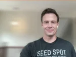 Smiling man with short brown hair on a webcam call, wearing a dark Seed Spot t-shirt.