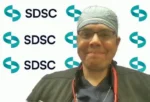 Healthcare professional wearing scrubs and a surgical cap, standing in front of an SDSC branded backdrop and smiling lightly.