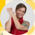 Portrait of a smiling woman with short brown hair, a red sleeveless top, visible arm tattoos, and hands clasped together.
