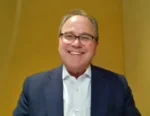 Smiling man in a dark suit and white shirt, wearing glasses, posing for a headshot against a mustard-yellow background.