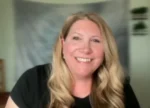 Portrait of a smiling woman with long blonde hair wearing a black top, indoors in a bright room.