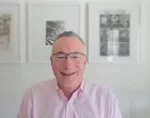 Smiling man with glasses in a light pink button-down shirt, seated indoors with framed artwork on the wall behind him.