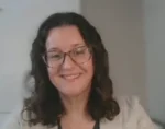 Smiling woman with shoulder-length dark hair and glasses, indoors in a light blazer.