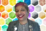 Smiling woman in a blazer with pins, posing in front of a colorful honeycomb background.