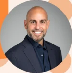 Professional head-and-shoulders portrait of a smiling bald man with a beard in a navy suit and shirt, against a gray backdrop with orange decorative shapes.