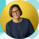 Smiling woman with short brown hair and glasses, wearing a blue blouse, posed in front of a bright yellow circle backdrop.